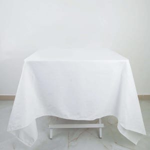 May include: A white tablecloth draped over a small, white table. The cloth is smooth and covers the entire table, with the edges hanging down. The background is a plain white wall and a marble floor.