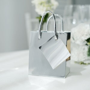 May include: A small, shiny silver gift bag with matching silver handles and a blank tag. The bag is square-shaped and reflects light, creating a bright and elegant appearance. Perfect for small gifts or party favors.