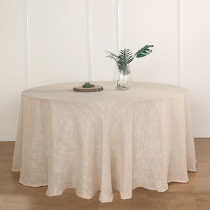May include: A round, beige linen tablecloth with a subtle textured pattern. The tablecloth is draped over a round table and has a slight drape. A small wooden tray with a green plant is on the table, along with a clear glass vase with green leaves.