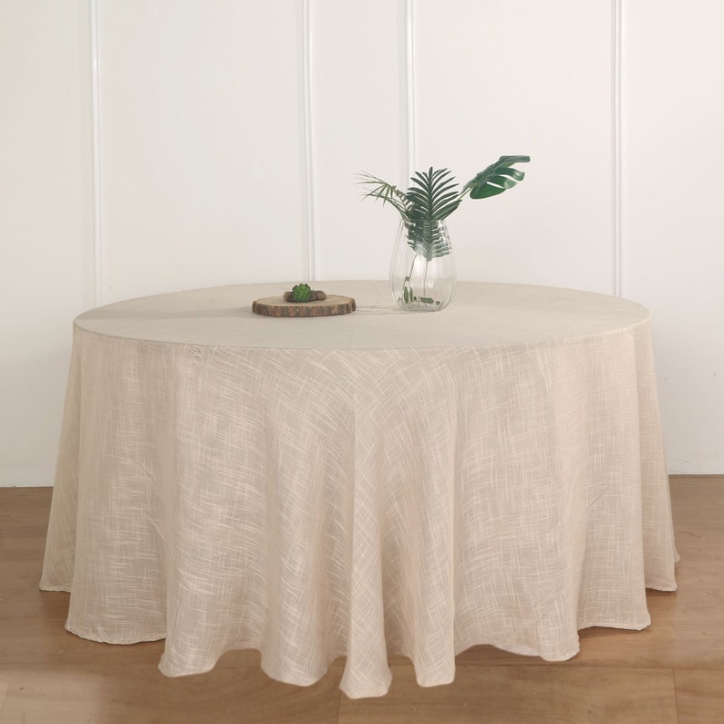 May include: A round, beige linen tablecloth with a subtle textured pattern. The tablecloth is draped over a round table and has a slight drape. A small wooden tray with a green plant is on the table, along with a clear glass vase with green leaves.