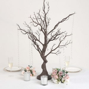 May include: A brown twig tree centerpiece with crystal strands hanging from the branches. Two bouquets of pink and white flowers are placed on either side of the tree. Two clear glass candle holders are on the table in front of the tree.