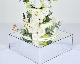 May include: A two-tiered white wedding cake decorated with gold flecks and topped with white roses and greenery. The cake is displayed on a square, mirrored cake stand.