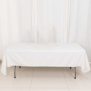 May include: A white rectangular tablecloth draped over a folding table with silver legs. The table is set against a white curtain backdrop, creating a clean and simple aesthetic. The table is approximately 60 inches wide and 120 inches long.
