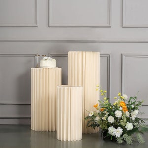 May include: Three white, fluted, round cake stands of different sizes. The stands are arranged in a row from largest to smallest. The largest stand is in the back, the middle stand is in the middle, and the smallest stand is in the front. The stands are on a gray background.