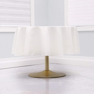 May include: A white round tablecloth draped over a gold round table base. The tablecloth is smooth and wrinkle-free.