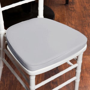 May include: A light gray chair cushion on a white chair. The cushion is square with rounded corners and a thin, darker gray border. The chair has a bamboo-style design.
