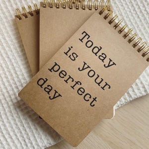 Könnte beinhalten: Drei spiralgebundene Notizbücher mit Kraftpapier-Einbänden und goldfarbener Drahtbindung. Das oberste Notizbuch trägt den Text "Today is your perfect day" in Schwarz. Die Notizbücher liegen auf einer weißen, strukturierten Oberfläche.