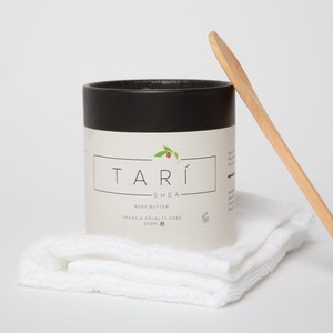 Könnte beinhalten: Ein weißes Glas mit schwarzem Deckel und einem Holzlöffel. Das Glas ist mit "TARI SHEA BODY BUTTER VEGAN & CRUELTY-FREE 250ml e" beschriftet. Das Glas steht auf einem weißen Handtuch.