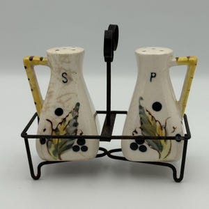 May include: A vintage salt and pepper shaker set. The ceramic shakers are off-white with a leaf and berry design. The handles are yellow with brown spots. The letters "S" and "P" are on the shakers. The set sits in a black metal holder.