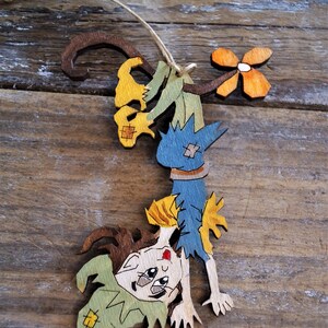 May include: A wooden ornament of a scarecrow with a blue and yellow shirt, brown hair, and a green hat. The scarecrow is hanging upside down from a branch with a yellow flower.