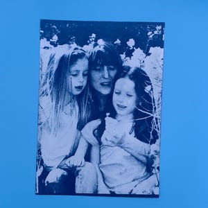 Peut inclure: Photographie en noir et blanc de trois personnes, probablement une m&egrave;re et deux enfants, en ext&eacute;rieur. L'image est imprim&eacute;e sur un papier rectangulaire, avec un fond bleu.