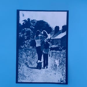 Peut inclure: Photographie en noir et blanc d'une famille se promenant sur un sentier. Une personne porte un enfant, tandis qu'un autre marche devant. Une maison et des arbres sont visibles en arri&egrave;re-plan.