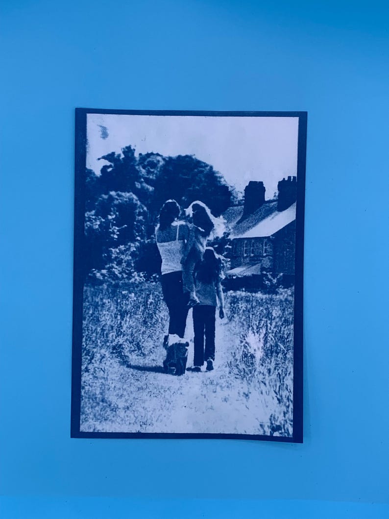 Peut inclure: Photographie en noir et blanc d'une famille se promenant sur un sentier. Une personne porte un enfant, tandis qu'un autre marche devant. Une maison et des arbres sont visibles en arri&egrave;re-plan.