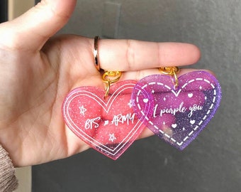 May include: Two heart-shaped keychains with glitter embedded in the clear plastic. One keychain is pink and says "BTS ARMY" in white lettering. The other keychain is purple and says "I purple you" in white lettering.
