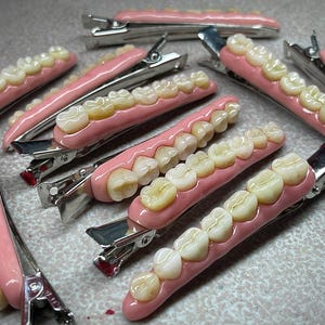 Puede incluir: Una colección de pinzas para el cabello. Cada pinza presenta una fila de dientes de aspecto realista colocados en una base rosa similar a la encía. Las pinzas son de metal y están dispuestas sobre una superficie texturizada.