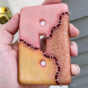 May include: A rectangular light switch cover plate featuring a textured, multi-colored surface that mimics skin. The design includes sections of pink, tan, and brown tones joined by dark red stitching and silver metal staples across the surface.