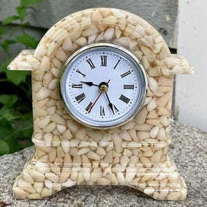 May include: A decorative table clock with a white clock face and black Roman numerals. The clock is encased in a beige material with a pattern of small, light-colored shapes. The clock has a silver-colored bezel and black clock hands.