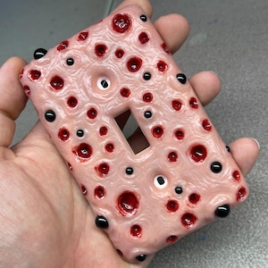 May include: A rectangular light switch cover plate with a textured, flesh-toned surface featuring numerous deep red, crater-like indentations and small, glossy black spherical protrusions scattered across the entire front face.
