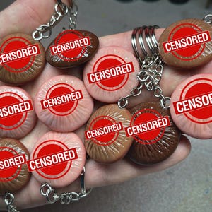 Peut inclure: Une collection de porte-clés avec des breloques rondes et texturées de différentes couleurs de peau. Chaque breloque porte un tampon rouge "CENSORED". Les porte-clés sont tenus dans une main.