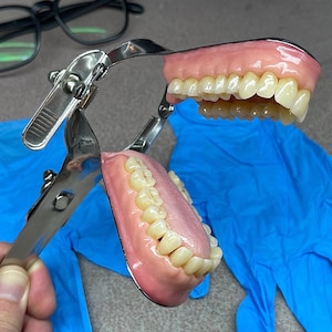 May include: A dental articulator holding a set of upper and lower dentures with pink acrylic gums and off-white teeth. The metal device features a hinged mechanism with a locking lever, designed to simulate jaw movement for dental modelling purposes.