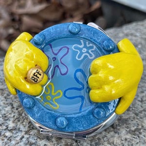 May include: A round, blue compact mirror with a cartoon design. The mirror features a blue background with white and purple starfish designs. Yellow, cartoon-style hands hold the mirror, and one holds a gold ring with the letters "BFF."