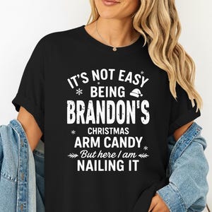May include: Black t-shirt with white text that reads "IT'S NOT EASY BEING BRANDON'S CHRISTMAS ARM CANDY But here I am NAILING IT." The shirt is worn with a denim jacket and shorts.
