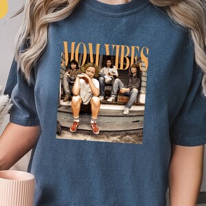 Puede incluir: Una camiseta azul marino con un estampado gr&aacute;fico de un grupo de personas sentadas en un porche. El gr&aacute;fico incluye el texto "MOM VIBES".