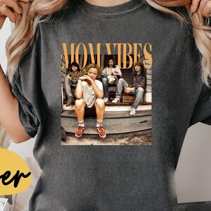 Puede incluir: Una camiseta gris oscuro con el texto "MOM VIBES" en letras naranjas. La camiseta presenta una foto de cuatro personas sentadas en un porche.