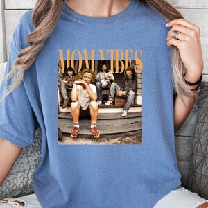 Puede incluir: Una camiseta azul con un estampado gr&aacute;fico de cuatro personas sentadas en un porche. El texto "MOM VIBES" est&aacute; impreso en naranja sobre la imagen.