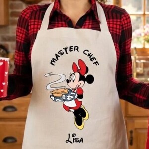 May include: Beige apron with a Minnie Mouse graphic holding a pie, the words "MASTER CHEF" and the name "LISA". The apron is worn over a red and black plaid shirt.
