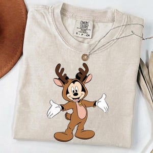 Puede incluir: Una camiseta beige de manga corta con un gráfico de un ratón de dibujos animados vestido con un disfraz de reno marrón con astas. La etiqueta interior dice Comfort Colors, 100% cotton, S-P-CH. Un collar de cadena plateada descansa sobre la tela.