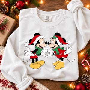 Puede incluir: Una sudadera blanca de cuello redondo con un gráfico de Mickey y Minnie Mouse con gorros de Papá Noel rojos y bufandas verdes. Los personajes están frente a frente y forman un corazón con las manos. Se ve una etiqueta de Comfort Colors.