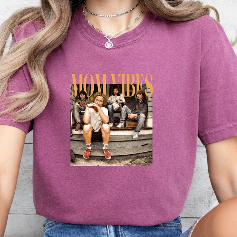Puede incluir: Una camiseta morada con un estampado gr&aacute;fico de un grupo de personas sentadas en un porche. El texto "MOM VIBES" est&aacute; impreso en naranja sobre la imagen.