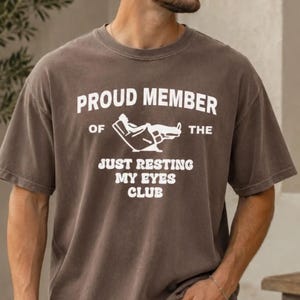Puede incluir: Camiseta marrón con el texto blanco "PROUD MEMBER OF THE JUST RESTING MY EYES CLUB". La camiseta presenta un gráfico de una persona reclinada en una silla. La camiseta tiene mangas cortas y cuello redondo.