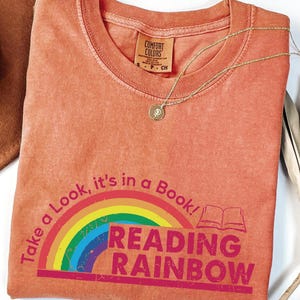 May include: A coral-colored t-shirt with the text "Take a Look, it's in a Book!" and "Reading Rainbow" in a pink font. A rainbow graphic is above the text. A gold necklace with a pendant is on the shirt.