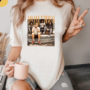Puede incluir: Camiseta blanca con un estampado gr&aacute;fico de un grupo de personas sentadas en un porche. El texto "MOM VIBES" est&aacute; impreso en naranja sobre la imagen.
