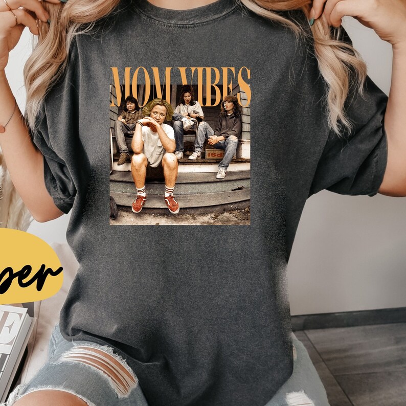 Puede incluir: Una camiseta gris oscuro con el texto "MOM VIBES" en letras naranjas. La camiseta presenta una foto de cuatro personas sentadas en un porche.