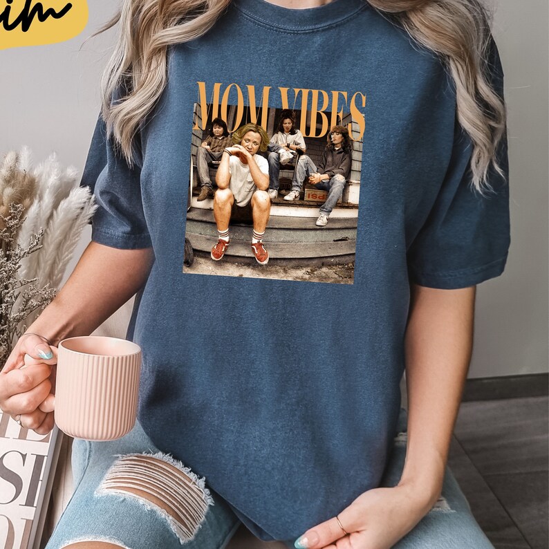 Puede incluir: Una camiseta azul marino con un estampado gr&aacute;fico de un grupo de personas sentadas en un porche. El gr&aacute;fico incluye el texto "MOM VIBES".