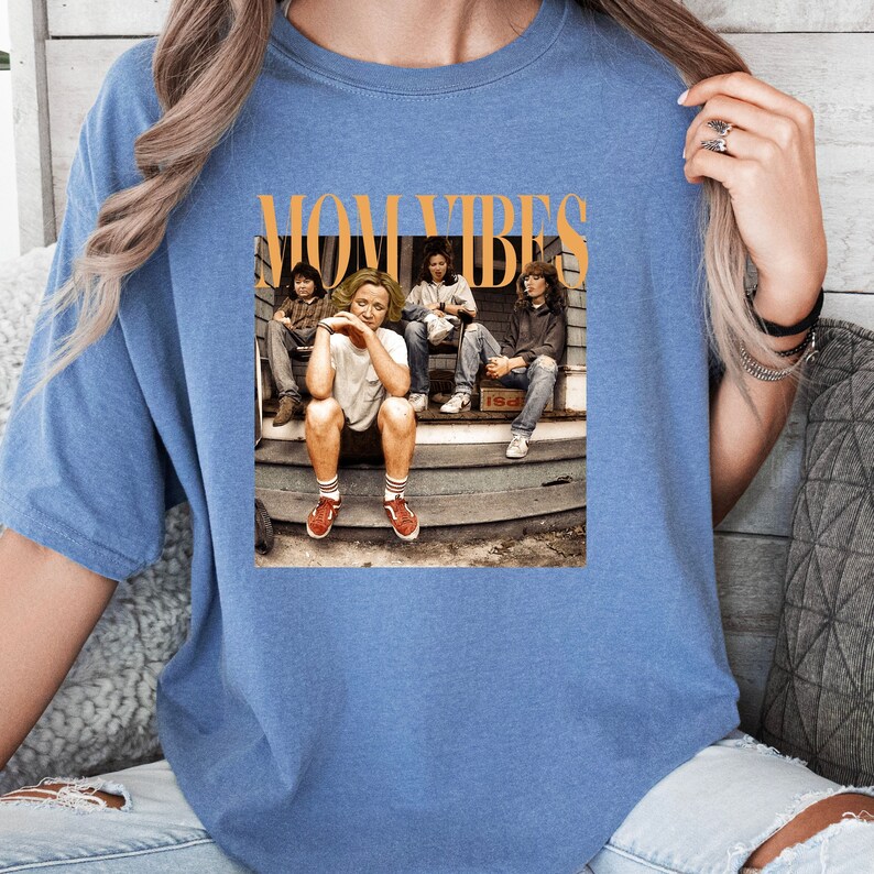 Puede incluir: Una camiseta azul con un estampado gr&aacute;fico de cuatro personas sentadas en un porche. El texto "MOM VIBES" est&aacute; impreso en naranja sobre la imagen.