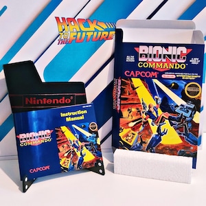 May include: A retro Bionic Commando video game box set. The blue box features action graphics and the game title. Includes an instruction manual and a black Nintendo game holder.