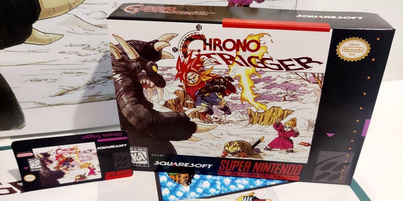 Puede incluir: Una copia en caja del juego de Super Nintendo "Chrono Trigger". La car&aacute;tula presenta una ilustraci&oacute;n din&aacute;mica de personajes en acci&oacute;n, con el t&iacute;tulo del juego en un lugar destacado. Tambi&eacute;n se ve una tarjeta m&aacute;s peque&ntilde;a con el t&iacute;tulo del juego.