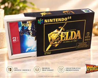 Peut inclure: Une boîte de jeu Nintendo 64 The Legend of Zelda Ocarina of Time noire avec un emblème d'épée et de bouclier doré. La boîte comprend un insert coulissant blanc contenant un boîtier de jeu Nintendo Switch avec une jaquette bleue et rouge.
