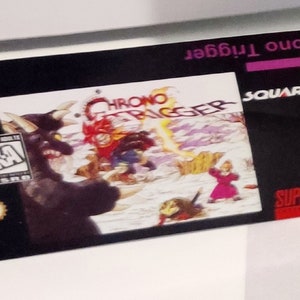 Puede incluir: Un cartucho de juego negro de Super Nintendo para Chrono Trigger. El cartucho presenta el t&iacute;tulo del juego, el logotipo de SquareSoft y una colorida ilustraci&oacute;n de personajes. El cartucho tiene licencia de Nintendo.