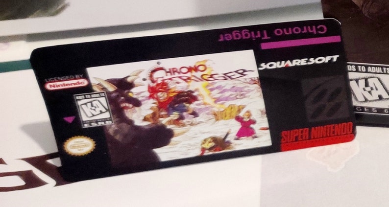 Puede incluir: Un cartucho de juego negro de Super Nintendo para Chrono Trigger. El cartucho presenta el t&iacute;tulo del juego, el logotipo de SquareSoft y una colorida ilustraci&oacute;n de personajes. El cartucho tiene licencia de Nintendo.