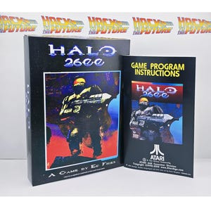 Puede incluir: Una caja de videojuego rectangular negra y un manual de instrucciones a juego para Halo 2600. La caja presenta un gráfico de un soldado sosteniendo un arma sobre un fondo rojo y azul con el texto Halo 2600 y A Game by Ed Fries.