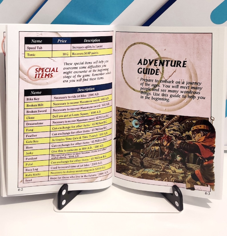Puede incluir: Un libro abierto sostenido por un atril negro. La p&aacute;gina izquierda presenta una lista de "Art&iacute;culos especiales" con detalles. La p&aacute;gina derecha muestra una "Gu&iacute;a de aventuras" con una ilustraci&oacute;n de una escena de batalla.