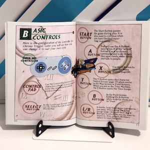 Puede incluir: Libro abierto con diagrama de mando de juego. La p&aacute;gina izquierda muestra un mando Super NES y el texto "Basic Controls". La p&aacute;gina derecha detalla las funciones de los botones como "Start", "A", "X", "Y", "B" y "L/R".