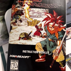 Puede incluir: Imagen del manual del juego de Super Nintendo "Chrono Trigger". La portada presenta personajes en un entorno de fantas&iacute;a, con el t&iacute;tulo del juego en primer plano. El manual es negro, con el logotipo de SquareSoft y una clasificaci&oacute;n "KA" para mayores de 6 a&ntilde;os.