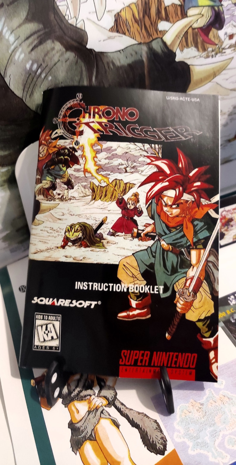 Puede incluir: Imagen del manual del juego de Super Nintendo "Chrono Trigger". La portada presenta personajes en un entorno de fantas&iacute;a, con el t&iacute;tulo del juego en primer plano. El manual es negro, con el logotipo de SquareSoft y una clasificaci&oacute;n "KA" para mayores de 6 a&ntilde;os.