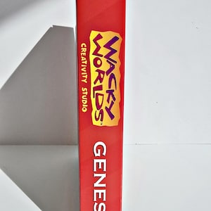 K&ouml;nnte beinhalten: Rote SEGA Genesis-Spielh&uuml;lle mit dem Spiel "Wacky Worlds Creativity Studio". Das Geh&auml;use zeigt das SEGA-Logo und das Wort "Genesis" vertikal. Der Spieletitel ist in Gelb und Lila.